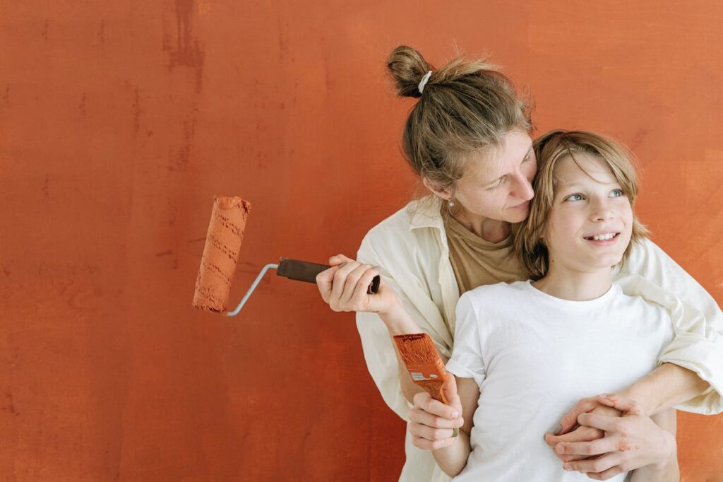 A woman hugging a boy holding a paint roller by an orange wall
